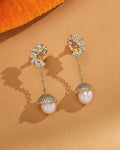 The Pearl Radiance CZ Jhumka by Chandrani Pearls features gold drop earrings with floral diamond-style tops, chain links, and round pearls in pave-set caps—festive jewelry elegantly displayed on a tan surface with an orange fabric accent.