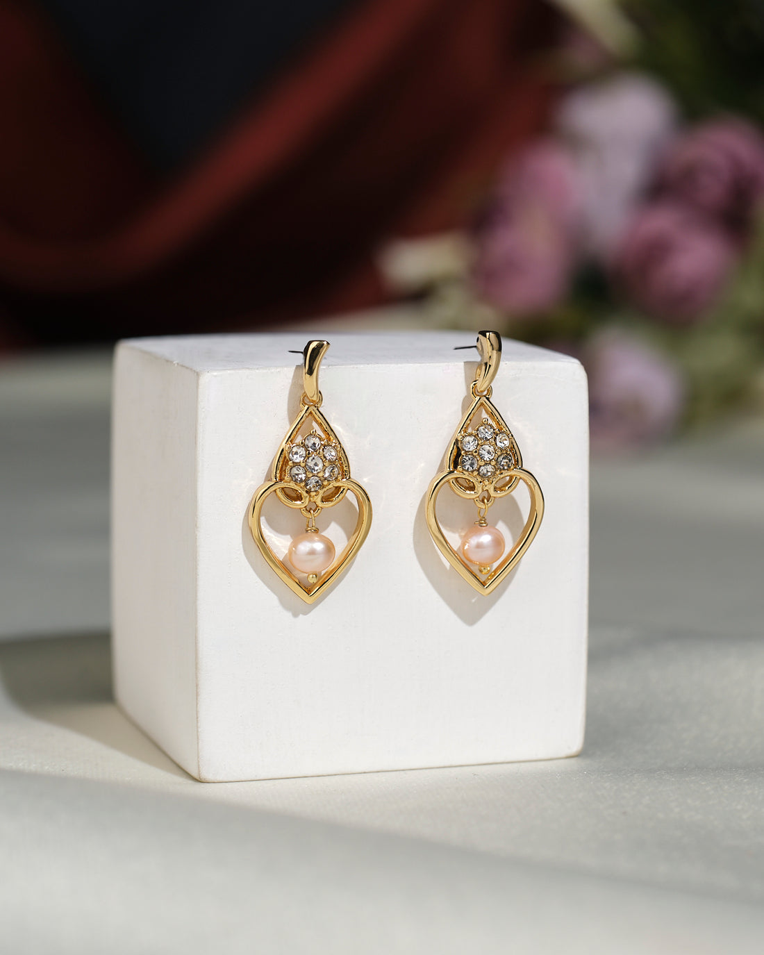 The Stylish Hang Earrings by Chandrani Pearls India feature gold heart-shaped designs with a polished finish, embellished with white stones and pink pearls, beautifully displayed on a white cube against a blurred floral backdrop.