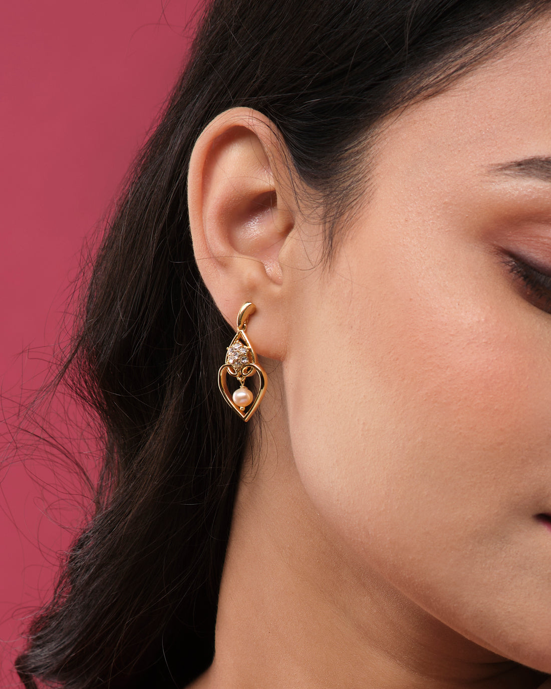 The Stylish Hang Earrings by Chandrani Pearls India feature gold heart-shaped designs with a polished finish, embellished with white stones and pink pearls, beautifully displayed on a white cube against a blurred floral backdrop.