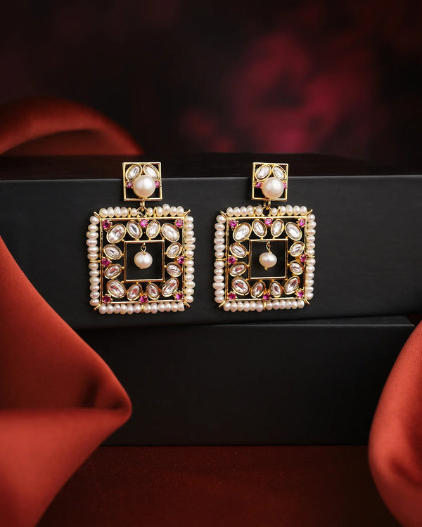 The Chantelle Pearl And Stone Chandeliers by Chandrani Pearls India are square gold-toned earrings embellished with pearls and gemstones, shown on a black box with red fabric details in the background.