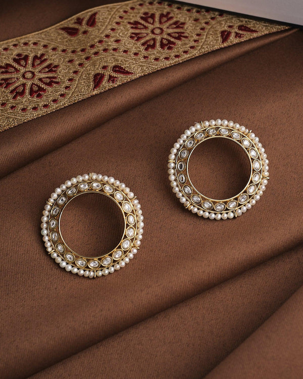 The Noor Classic Grace Studs by Chandrani Pearls—circular gold pearl earrings with white stones—embody quiet luxury and are displayed on brown fabric with an embroidered gold and red border.