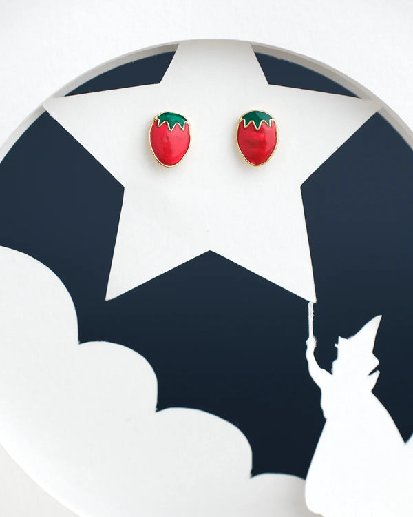 The Stawberry Stud Earring by Chandrani Pearls India features red strawberry-shaped earrings on a star-shaped background with a silhouette of a cat holding an umbrella.