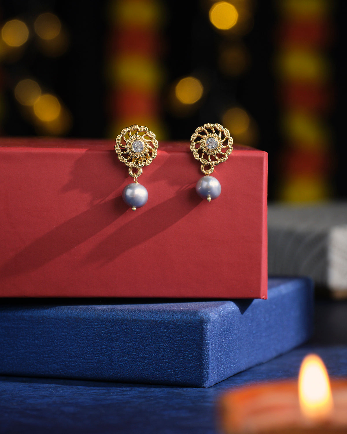 Jaffri Artworked Pearl Earrings by Chandrani Pearls showcased on a red box, accompanied by a blurred background and an illuminated candle in the foreground.