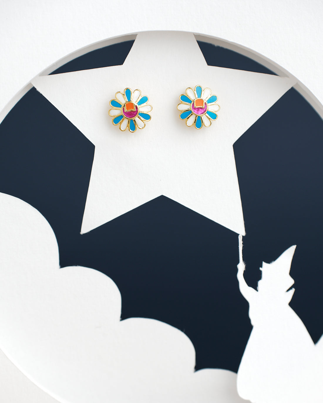 Chandrani Pearls India's Sunflower Stud Earrings, featuring two daisy-shaped designs with blue petals and red centers, are displayed on a circular background with a silhouette of a person pointing to a star.