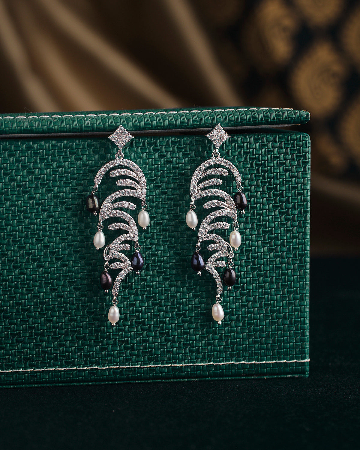 A pair of ornate earrings from Chandrani Pearls, named The Ratriratna-Symbolizing Purity, featuring silver design with pearls and black beads, are displayed on a textured green surface.