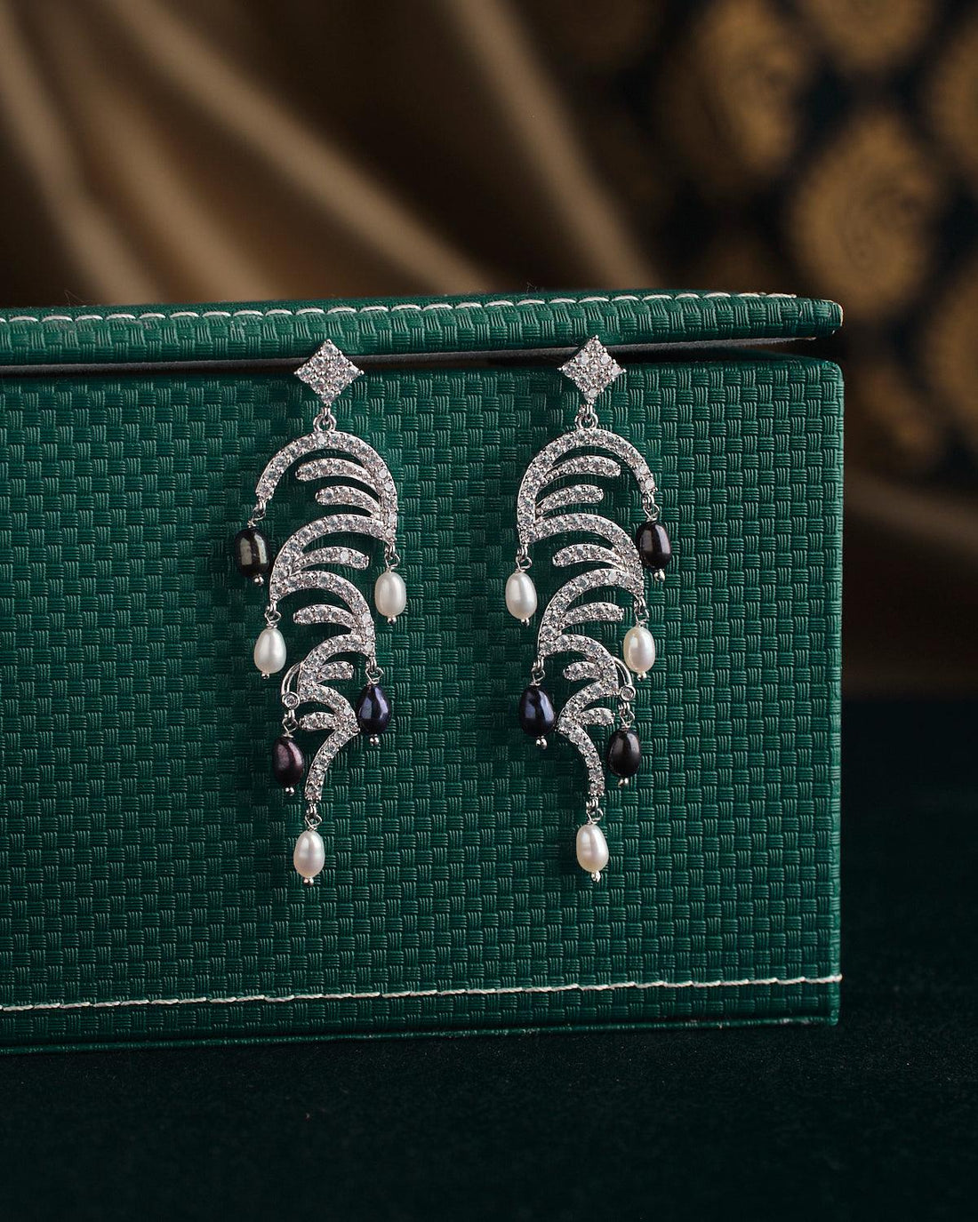 A pair of ornate earrings from Chandrani Pearls, named The Ratriratna-Symbolizing Purity, featuring silver design with pearls and black beads, are displayed on a textured green surface.