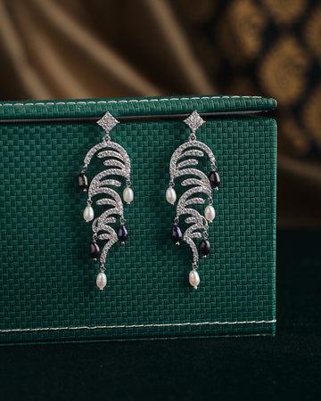 A pair of ornate earrings from Chandrani Pearls, named The Ratriratna-Symbolizing Purity, featuring silver design with pearls and black beads, are displayed on a textured green surface.