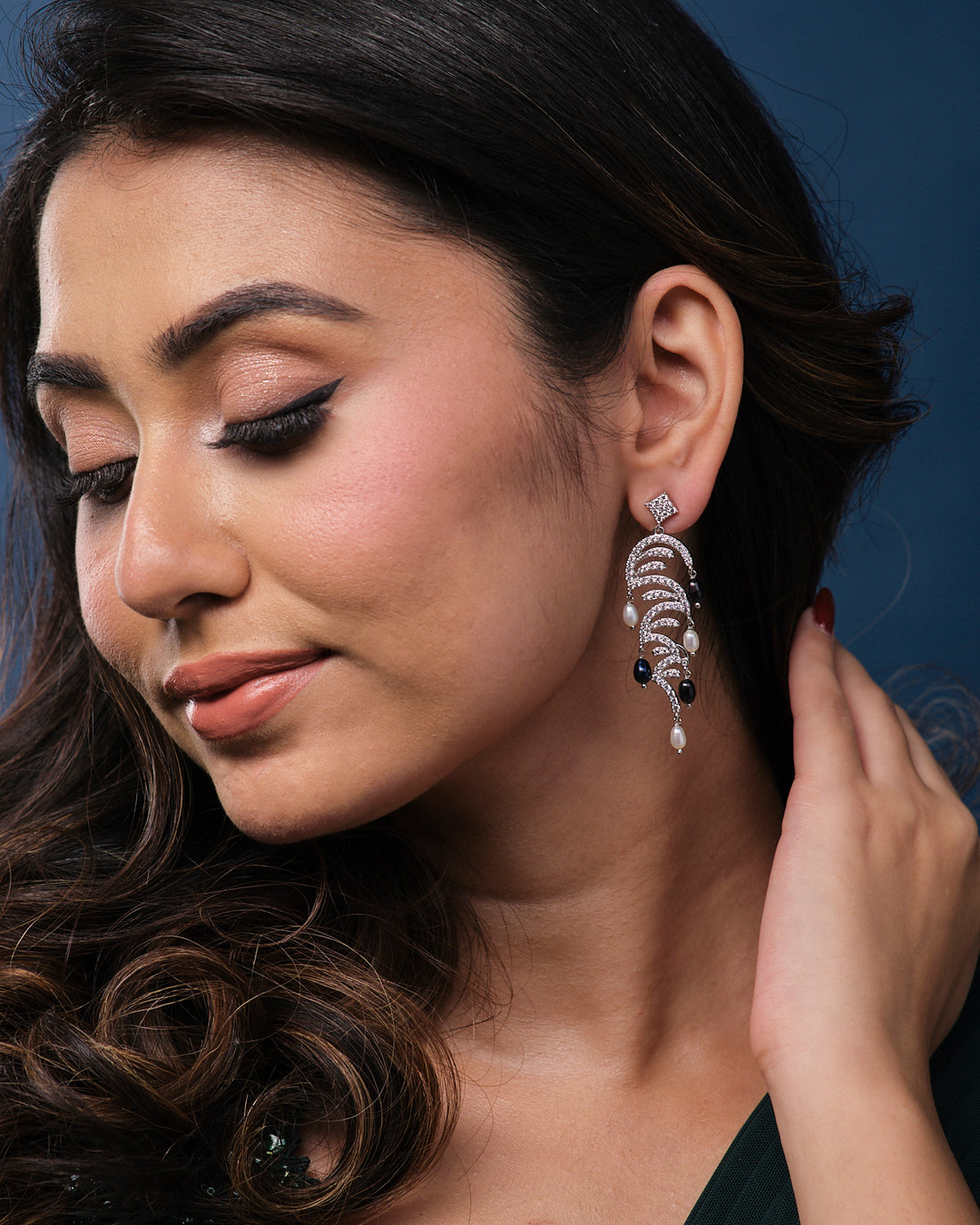 A pair of ornate earrings from Chandrani Pearls, named The Ratriratna-Symbolizing Purity, featuring silver design with pearls and black beads, are displayed on a textured green surface.