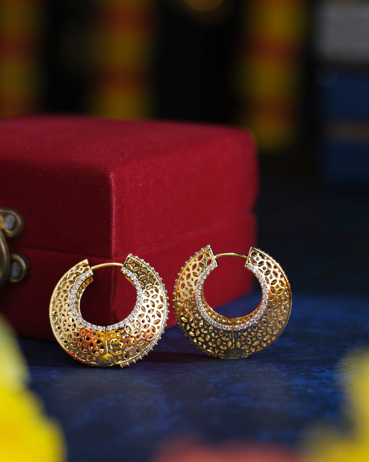 Two intricately designed Vivaciously Designed Huggie Silver Earrings from Chandrani Pearls in front of a red velvet box, placed on a dark blue surface.