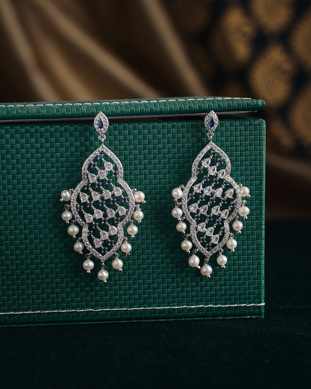 Navodha - The Wedded Dazzle Pearl Earrings by Chandrani Pearls, featuring diamond patterns and pearl accents, elegantly displayed on a textured green surface.