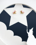 A pair of The Cute Foal Stud Earrings by Chandrani Pearls India with pink manes sits on a white star cutout against a dark background, beneath which a wizard silhouette points a wand.