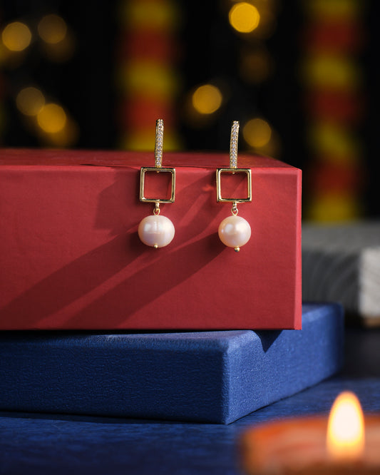 The Conifer Cone Pearl Earrings by Chandrani Pearls are elegantly displayed on a red box, set against a blurred festive backdrop adorned with bokeh lights. A small candle adds warmth to the foreground.