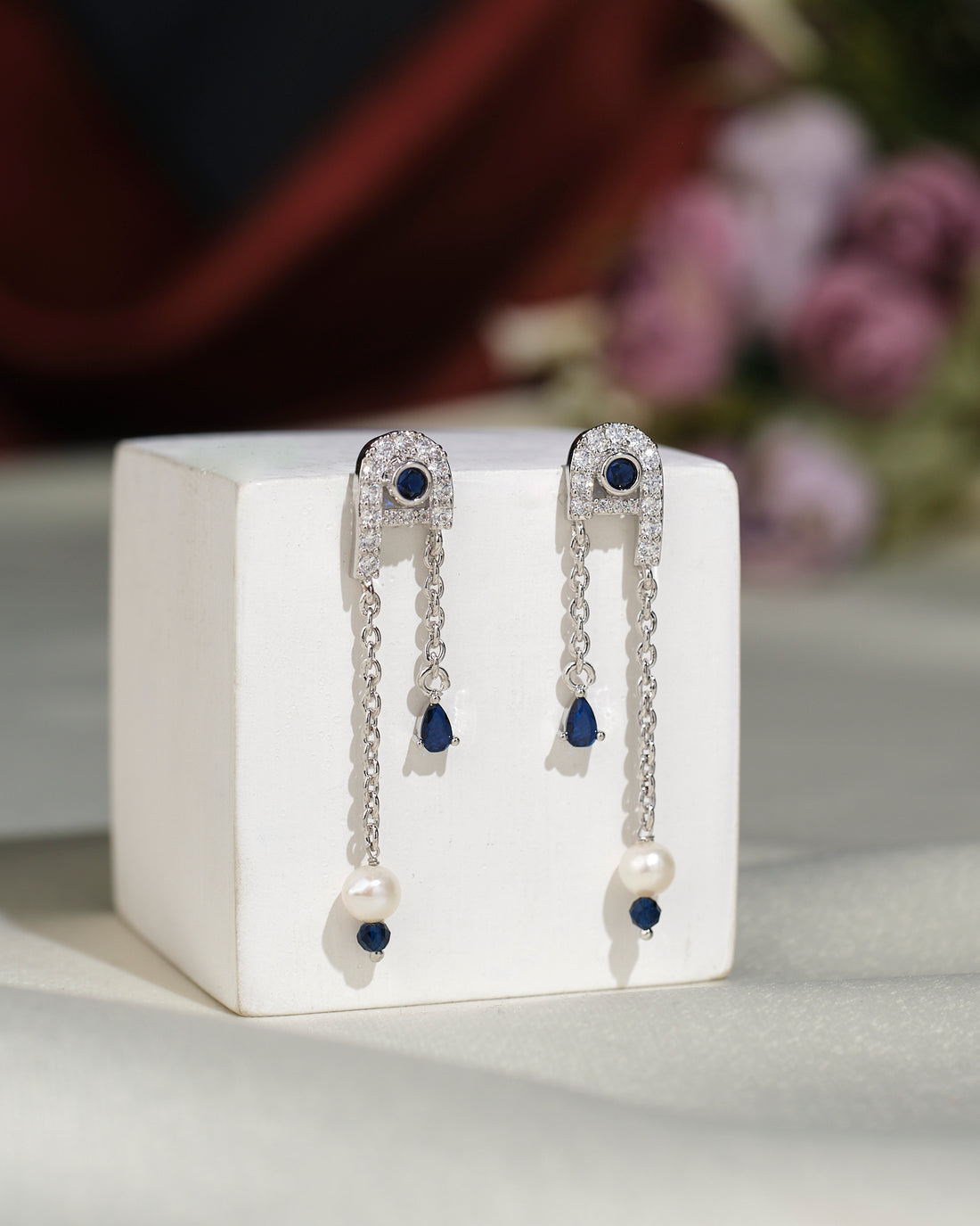 The Stylish Hang Earrings by Chandrani Pearls India feature silver drops with blue stones, pearls, and chain details, displayed upright on a white cube for subtle everyday glamour against a blurred floral background.