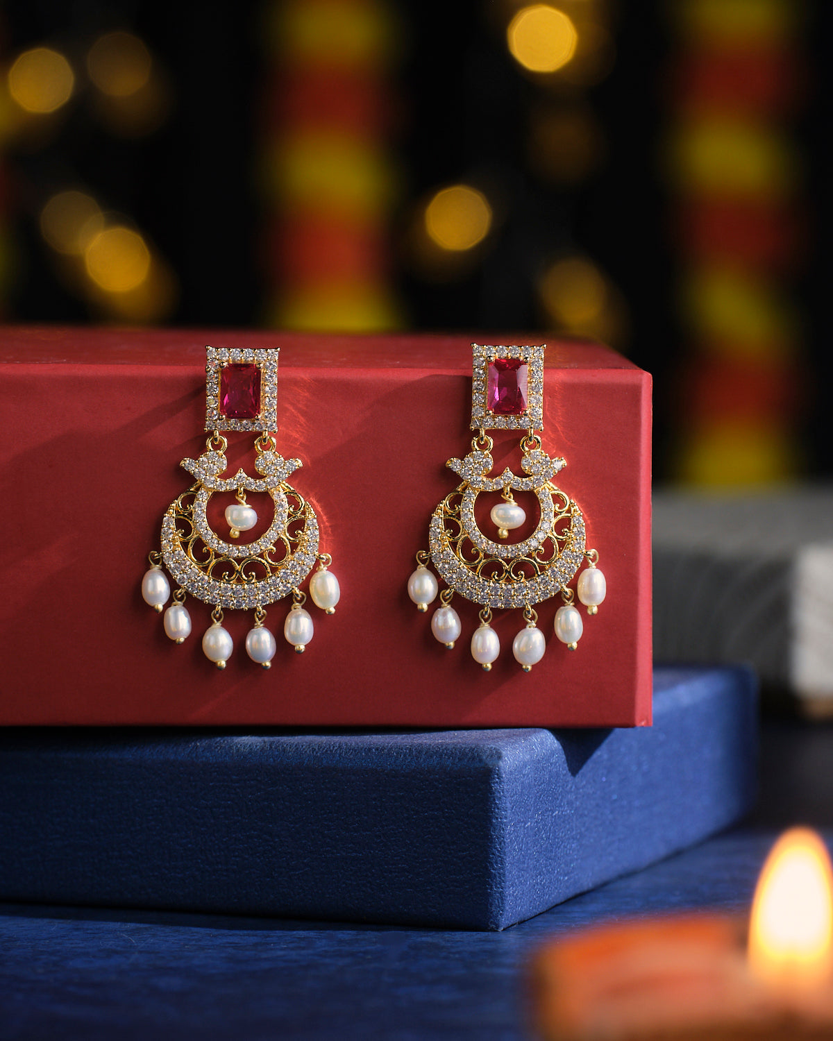 Chandrani Pearls' Sparkling Wreath Pearl Earrings, featuring dazzling red gemstones and pearls, are elegantly displayed on a red box with a blurred festive background.