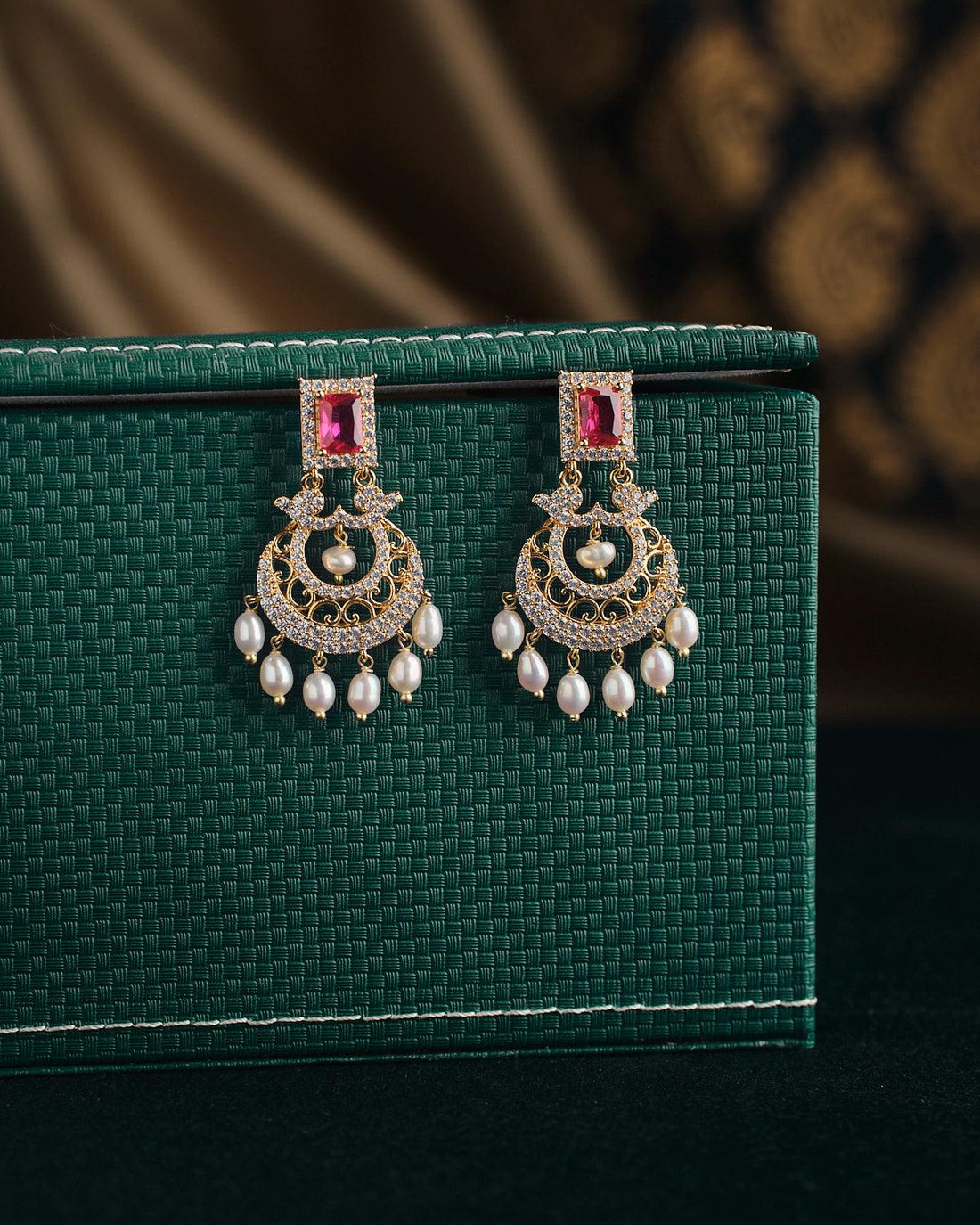 Gold ornate "Sparkling Wreath Pearl Earrings" by Chandrani Pearls, featuring red square stones and dangling pearls, are elegantly displayed on a green textured box.