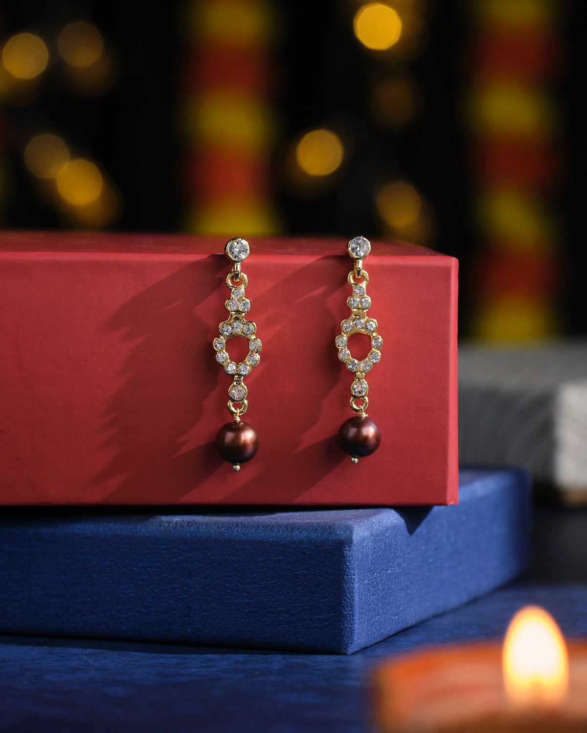 Radiant Tower Pearl Earrings by Chandrani Pearls showcased on red and blue boxes, with blurred lights creating a dazzling backdrop.