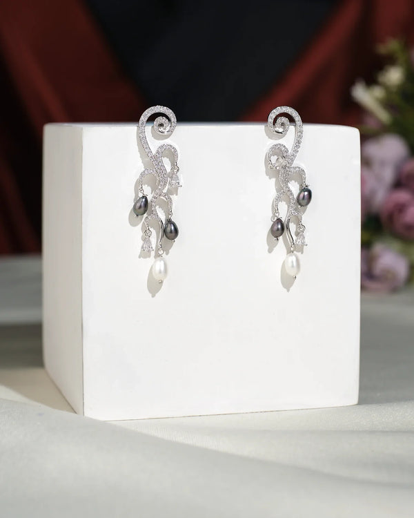Stylish Hang Earrings from Chandrani Pearls India feature swirling silver designs, sparkling gemstones, and dangling black and white pearls, exuding elegance as they are displayed on a white cube.