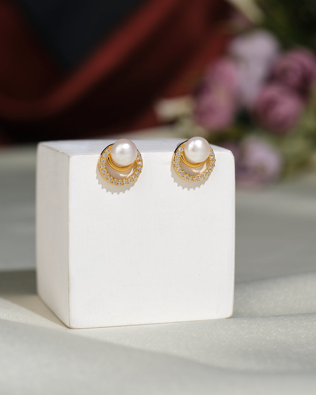 The Cute Stud Earrings by Chandrani Pearls India feature round pearls and small clear stones, offering classic elegance and style. They are displayed on a white block with a blurred background.
