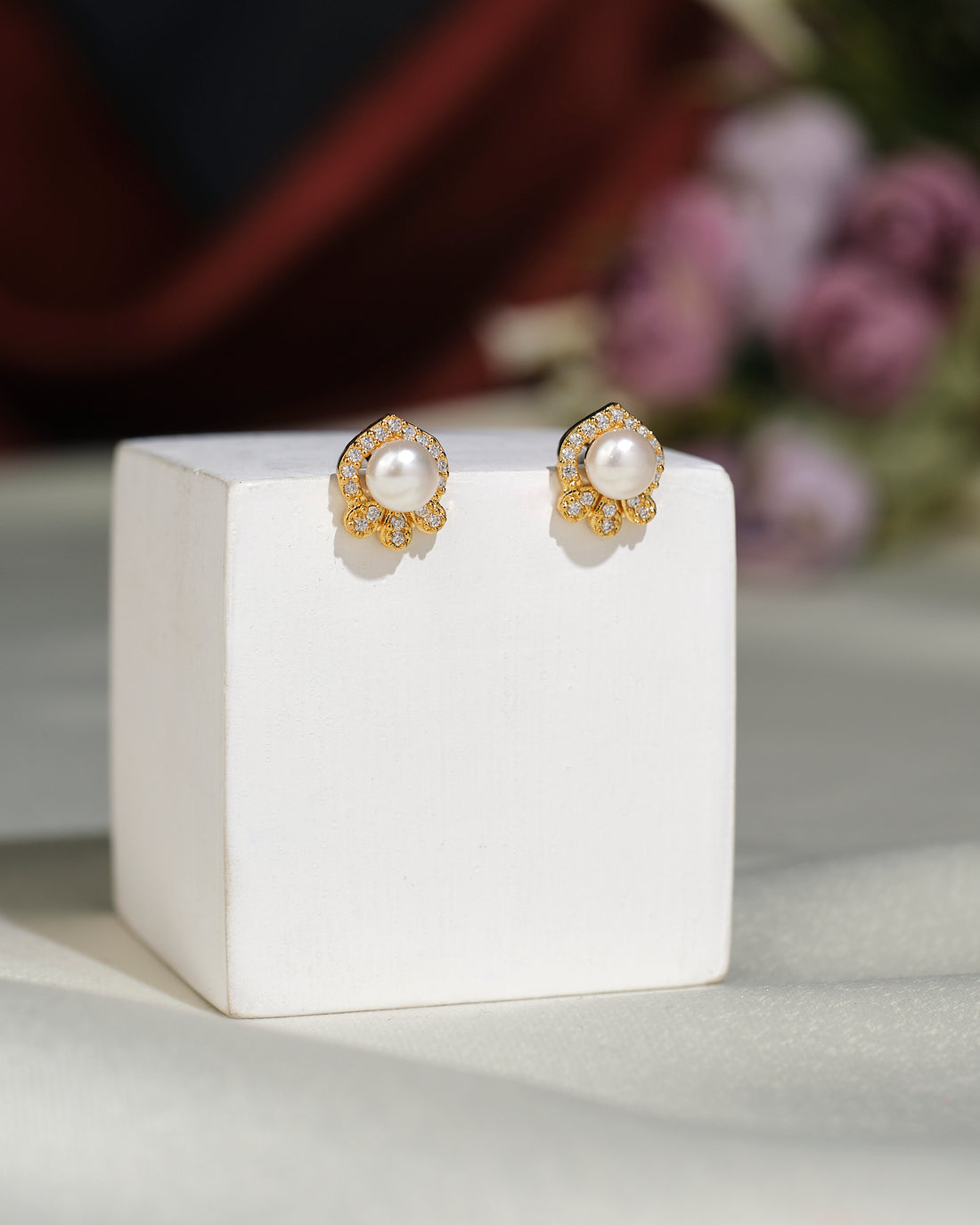 The Pretty Stud Earrings from Chandrani Pearls India, featuring gold and pearl studs with small clear gemstones, are displayed on a white stand with a blurred floral background, showcasing timeless elegance.