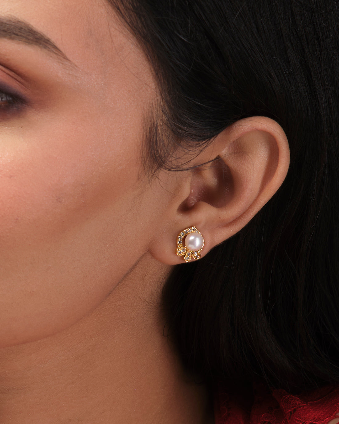The Pretty Stud Earrings from Chandrani Pearls India, featuring gold and pearl studs with small clear gemstones, are displayed on a white stand with a blurred floral background, showcasing timeless elegance.