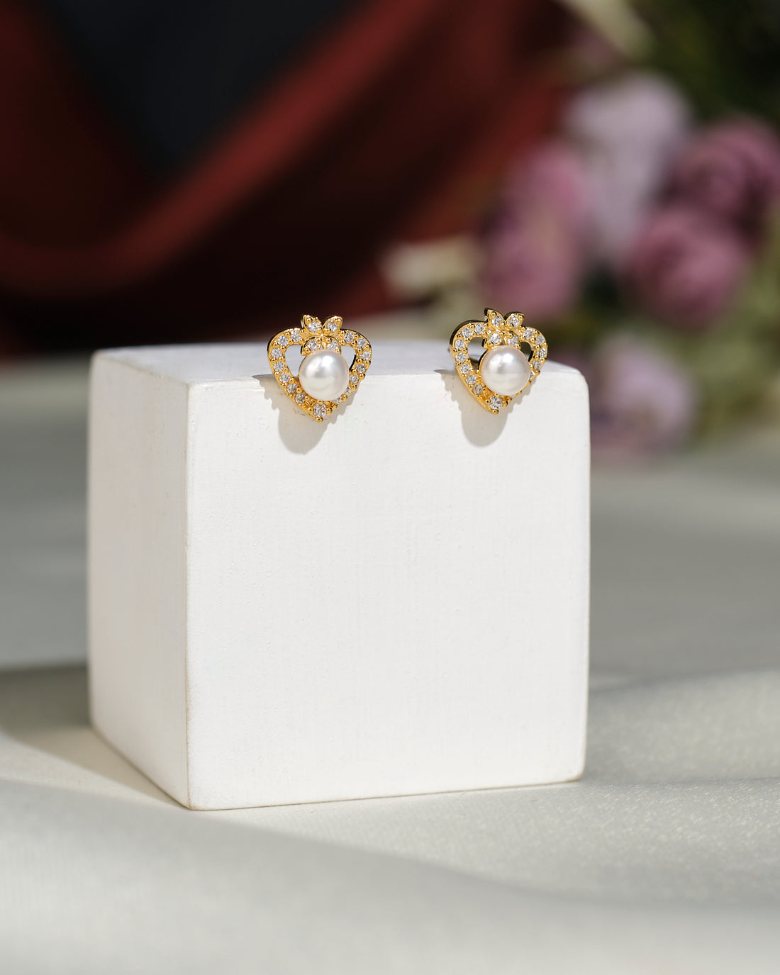 Cute Stud Earrings by Chandrani Pearls India feature gold heart-shaped studs with central pearls, offering timeless style. They are elegantly displayed on a white cube, set against a blurred floral background.
