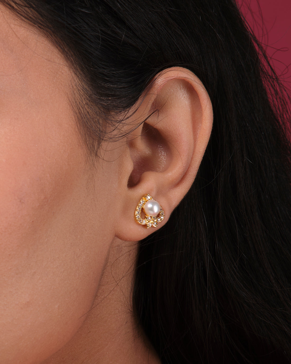 Cute Stud Earrings by Chandrani Pearls India feature gold heart-shaped studs with central pearls, offering timeless style. They are elegantly displayed on a white cube, set against a blurred floral background.