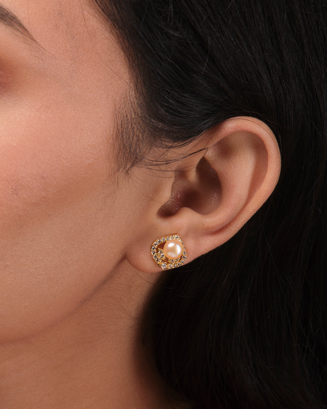 Simple Stud Earrings by Chandrani Pearls India, featuring gold and pearl accents, are displayed on a white square pedestal with blurred flowers in the background, highlighting their timeless sophistication.
