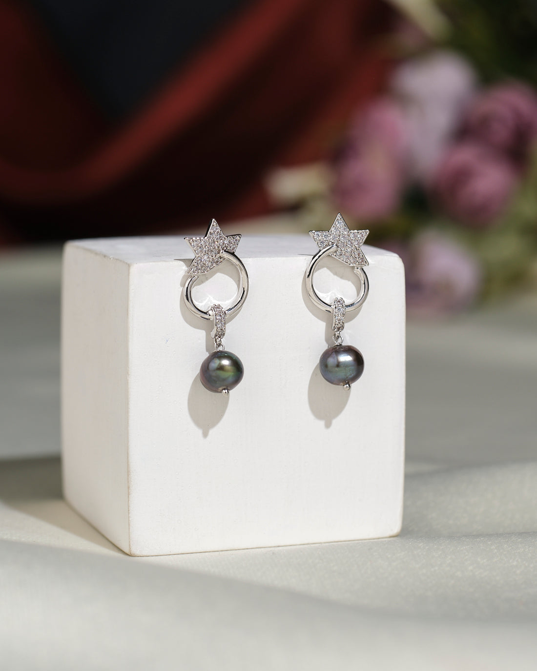 The Trendy CZ Hang Earrings by Chandrani Pearls India feature star-shaped silver designs with black pearls, displayed on a white stand against a blurred floral background—perfect for adding sophistication to any look.