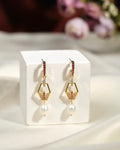 The Belinha Drop Earrings by Chandrani Pearls India feature gold geometric shapes, pink gemstones, and a pearl dangle, shown on a white cube against a soft, neutral fabric background.