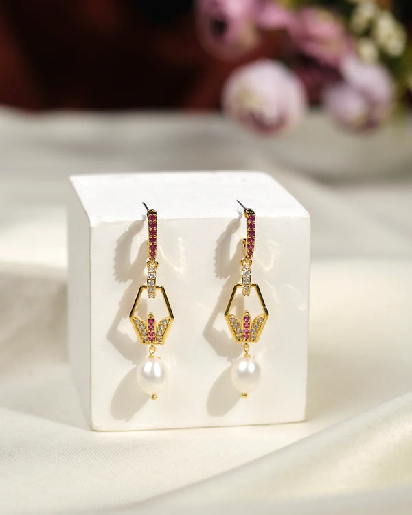 The Belinha Drop Earrings by Chandrani Pearls India feature gold geometric shapes, pink gemstones, and a pearl dangle, shown on a white cube against a soft, neutral fabric background.