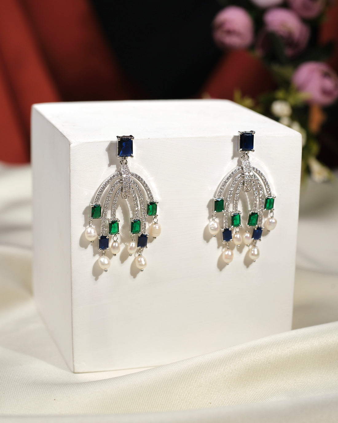 The Radiant Droplet Earrings by Chandrani Pearls India feature luxury silver with blue and green gemstones and delicate pearl drops, beautifully displayed on a white block—ideal for adding elegance to any outfit.