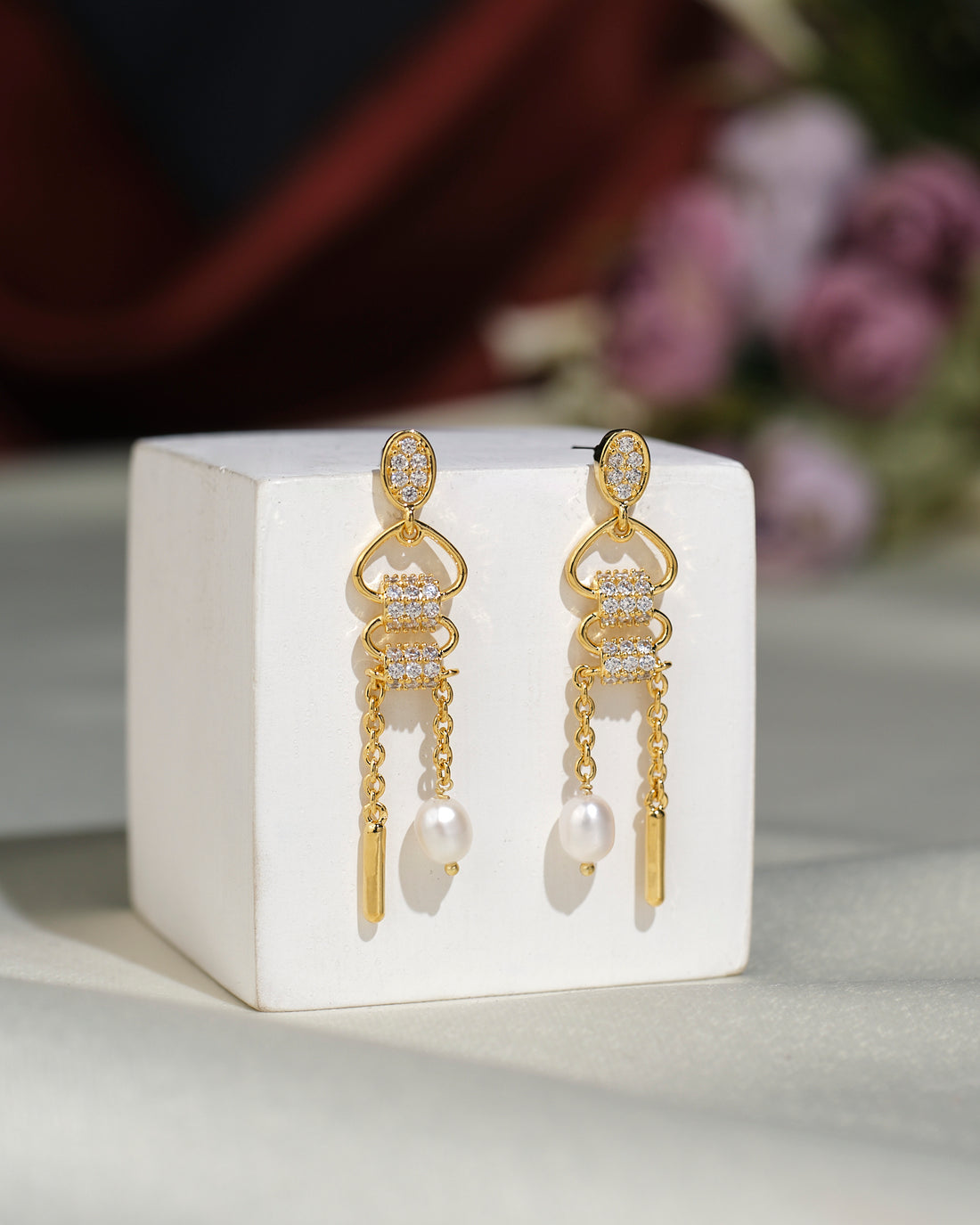 The Fashionable CZ Hang Earrings by Chandrani Pearls India feature lustrous clear stones, delicate chains, and a single dangling pearl, elegantly displayed on a white cube.