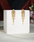 The Stylish Hang Earrings by Chandrani Pearls India feature gold dangles with pearl drops and rhinestone accents, elegantly displayed on a white cube against a softly blurred floral background.