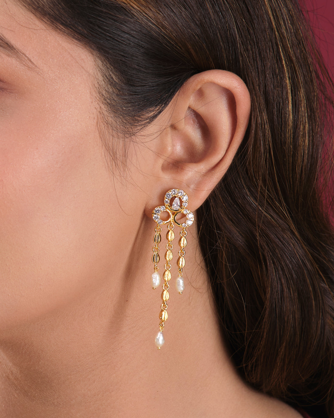 The Stylish Hang Earrings by Chandrani Pearls India feature gold dangles with pearl drops and rhinestone accents, elegantly displayed on a white cube against a softly blurred floral background.