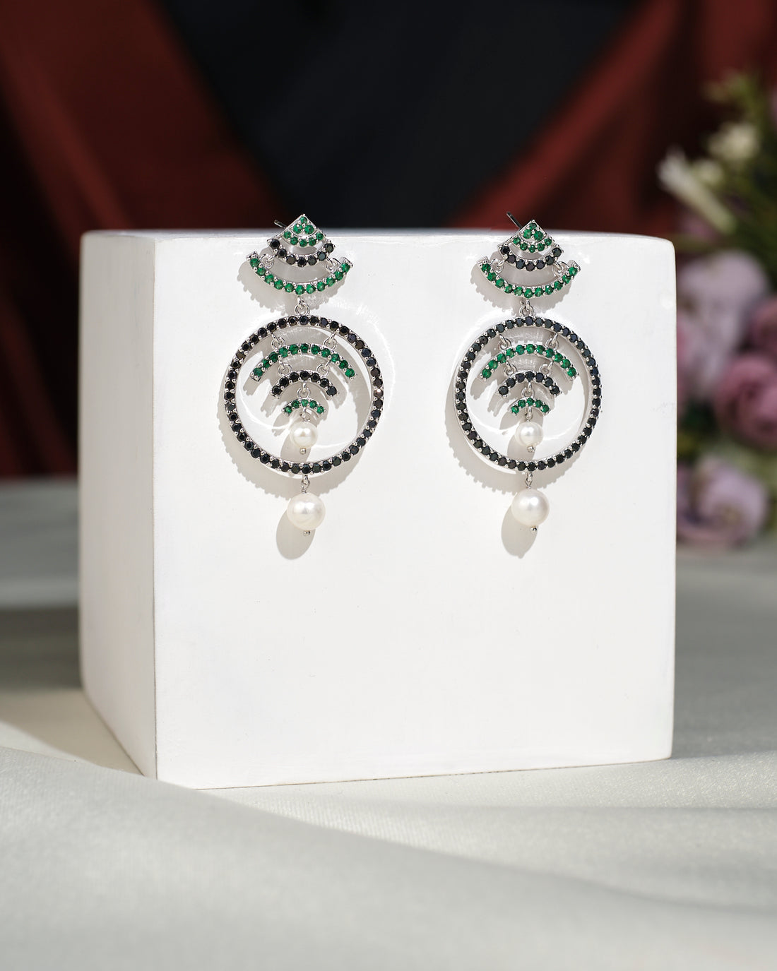 The Fashionable CZ Hang Earrings by Chandrani Pearls India feature green and black stones, circular designs, and elegant pearl drops, displayed on a white cube with a blurred floral background for timeless appeal.