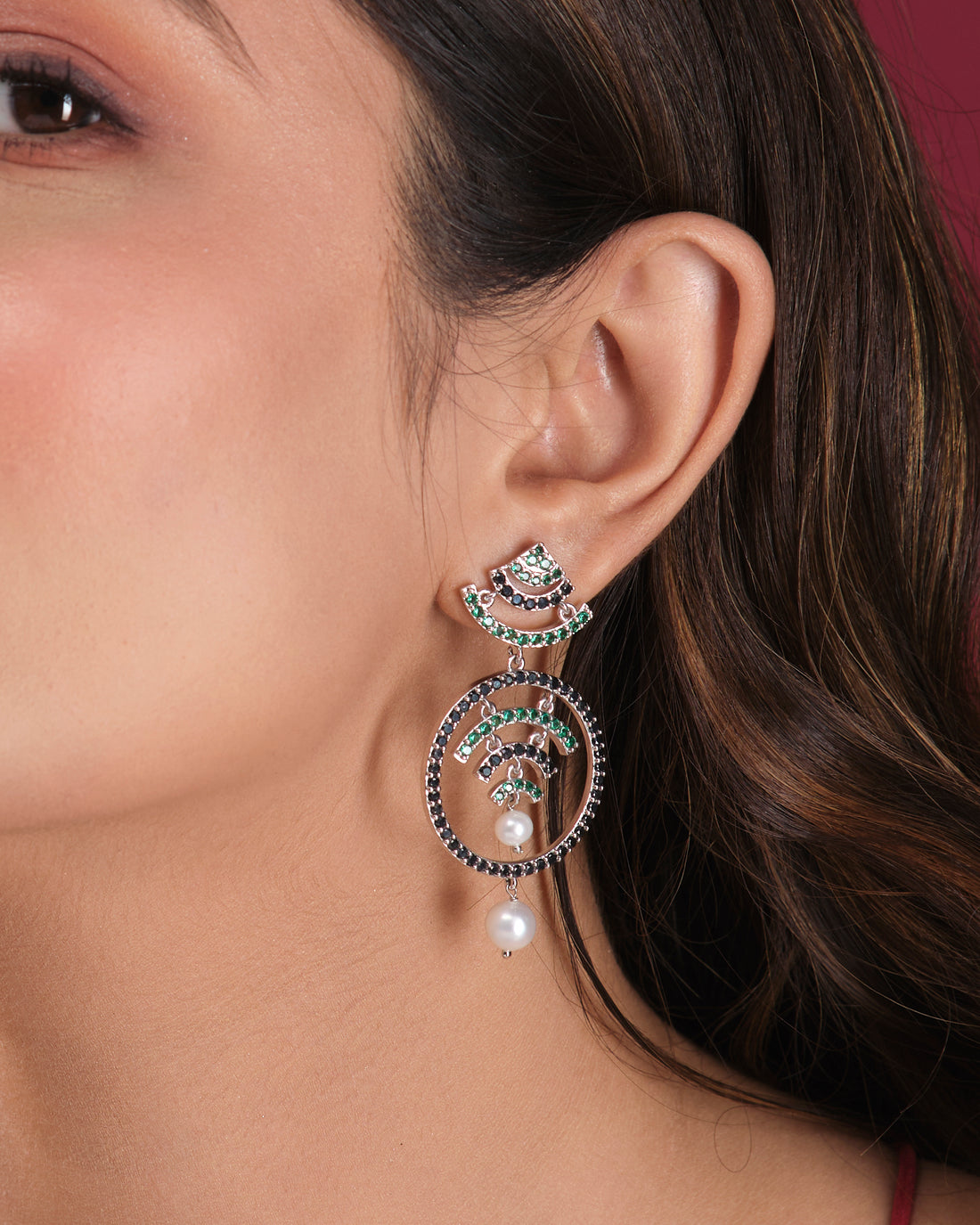 The Fashionable CZ Hang Earrings by Chandrani Pearls India feature green and black stones, circular designs, and elegant pearl drops, displayed on a white cube with a blurred floral background for timeless appeal.