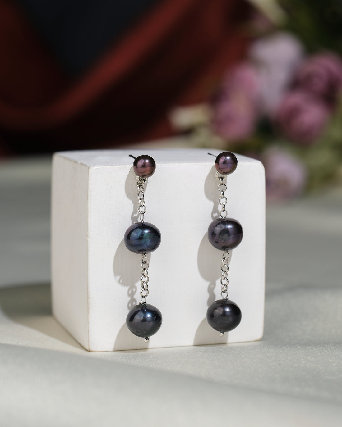 Chandrani Pearls India's Stylish Hang Pearl Earrings feature three black pearls on each earring, linked by silver chains. Displayed on a white block with a blurred floral background, they offer timeless elegance.