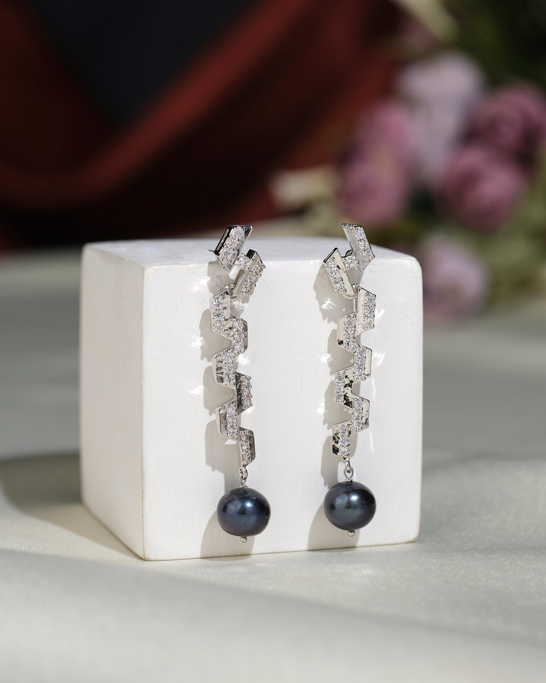 Fashionable CZ Hang Earrings by Chandrani Pearls India: chic long silver earrings with a zigzag design, sparkling stones, and round black pearls, elegantly displayed on a white block.