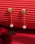 The Pretty Heart CZ 925 Silver Earrings by Chandrani Pearls India feature gold heart shapes with clear stones, chain links, and dangling white pearls, stylishly displayed against a red textured background.