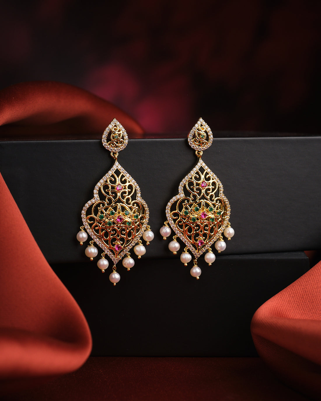 The Zuly Multi Stone Pearl Earrings by Chandrani Pearls feature ornate gold, colored enamel, and iridescent pearl dangles—ideal statement cocktail earrings—shown against a black box with red fabric accents.