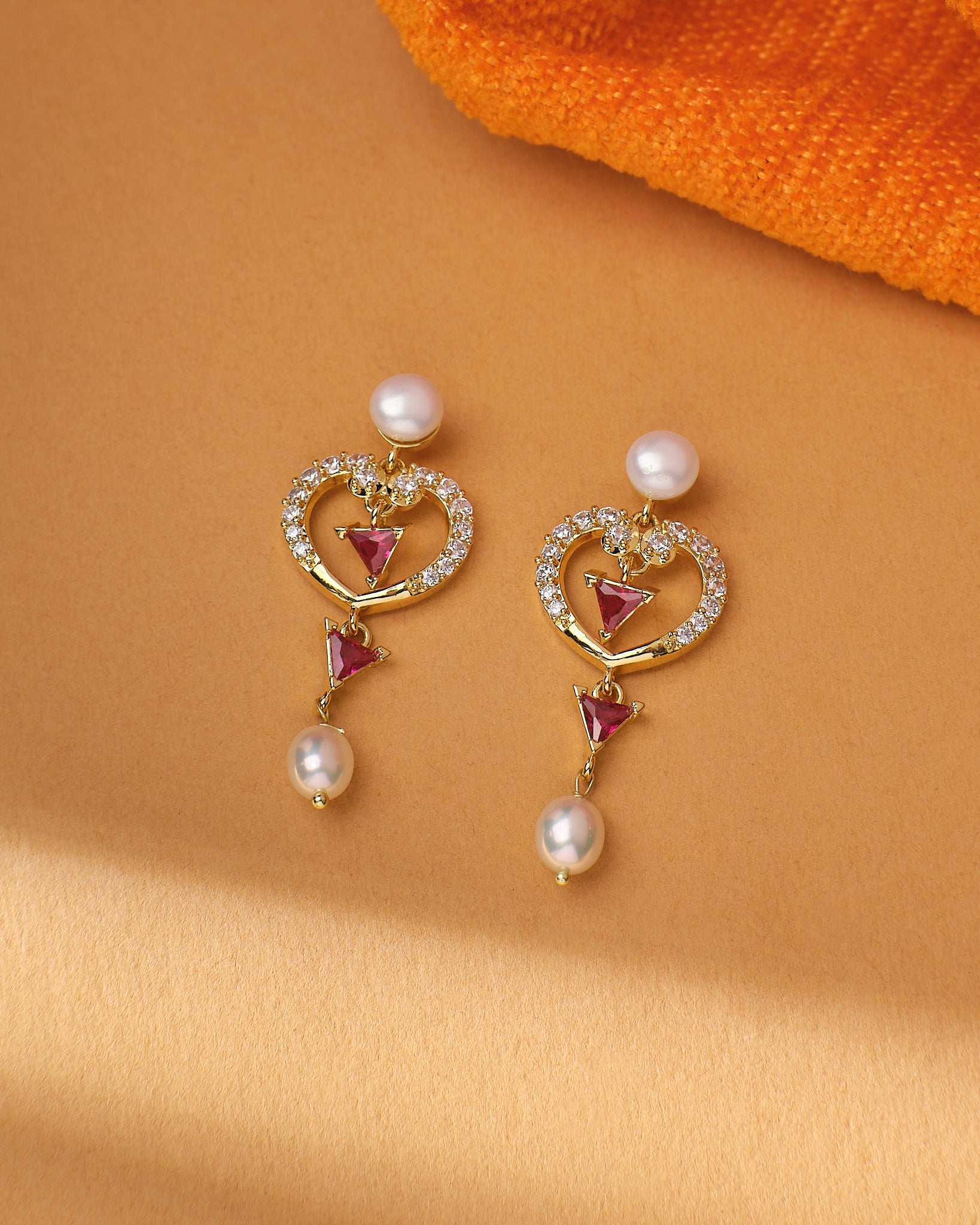 The Celestial Heart Glow CZ Pearl Earrings by Chandrani Pearls feature gold settings with heart and triangle red stones, small white pearls, and circular CZ stones, showcased on beige with a touch of orange fabric.