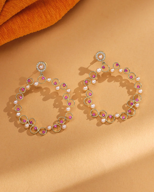 The Timeless Elegance Pearl CZ Jhumka by Chandrani Pearls features large circular gold jhumkas with pink gemstones, CZ stones, and natural pearls, displayed on a light brown surface with orange fabric in the corner.