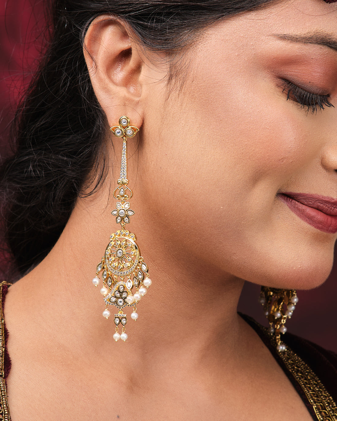 Dreamy Desire Pearl Earrings by Chandrani Pearls, featuring ornate gold and pearls with handcrafted twisted chains, are displayed on black boxes against a dark red satin background.
