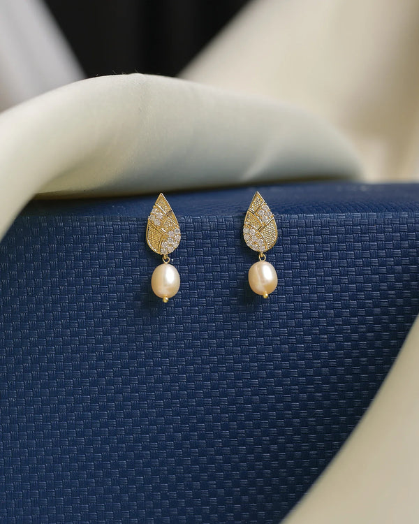 Chandrani Pearls’ earrings, crafted with high-shine CZ stones in gold leaf-shaped tops and smooth pearls, are displayed on a textured dark blue surface.