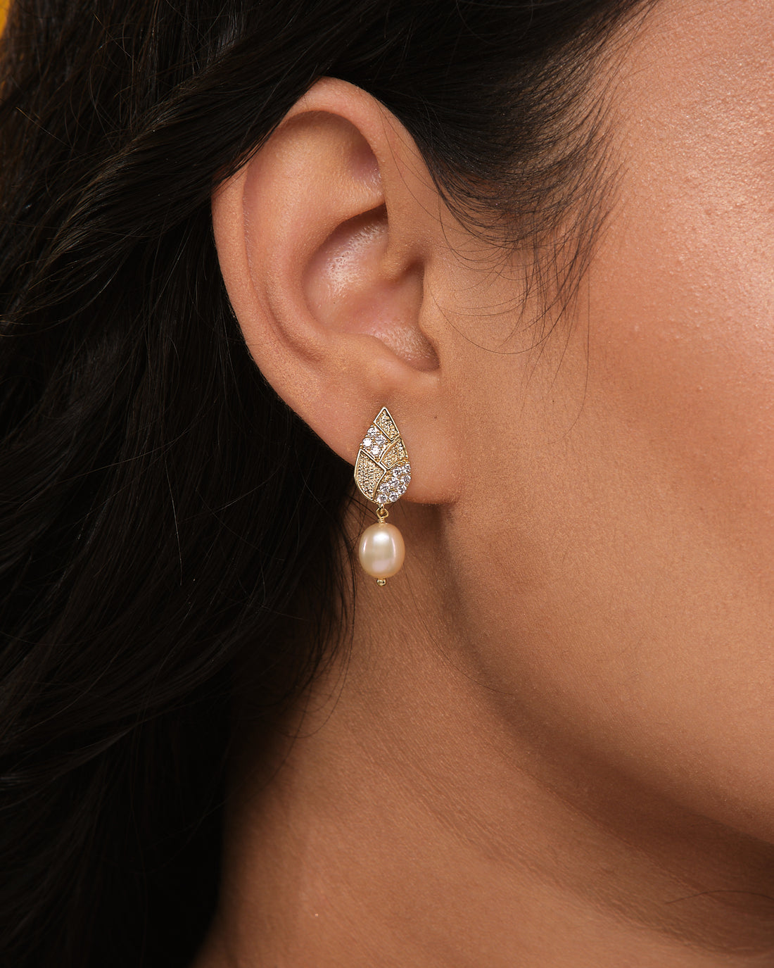Chandrani Pearls’ earrings, crafted with high-shine CZ stones in gold leaf-shaped tops and smooth pearls, are displayed on a textured dark blue surface.
