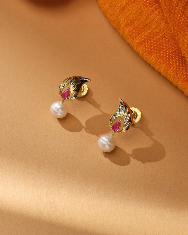 The Twilight Glow CZ Earrings by Chandrani Pearls feature gold with pearl highlights and red gemstone accents, displayed on beige with orange textured fabric—an elegant, twilight-inspired design for special evenings.