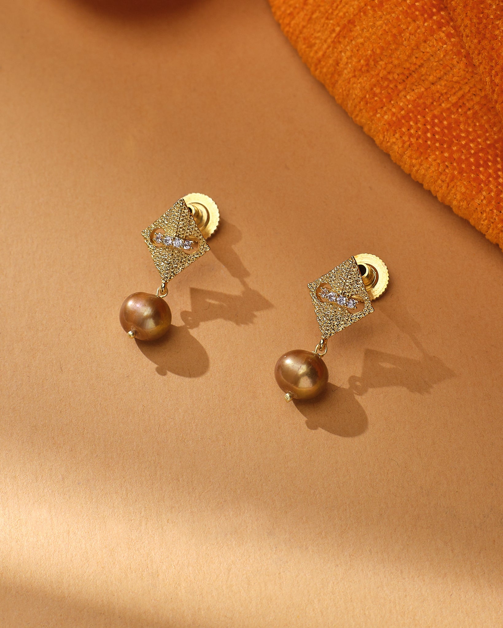 The Dainty Pearl CZ Earrings by Chandrani Pearls feature gold-toned diamond-shaped studs with clear stones and brown pearl jhumkas, shown on a beige surface with orange fabric for casual elegance.