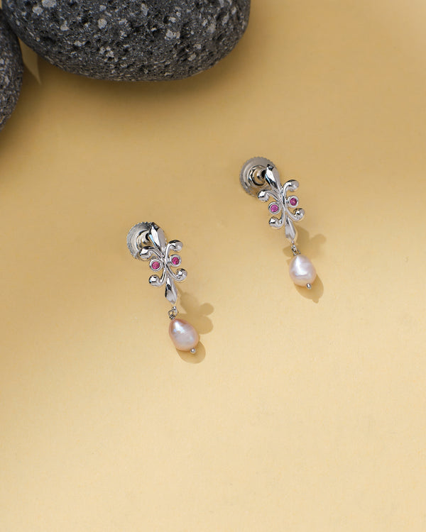 Chandrani Pearls Dangling Pearl Earrings feature delicate silver, pink gemstones, sparkling CZ stones, and elegant pearl drops, displayed on a beige surface with black stones nearby.