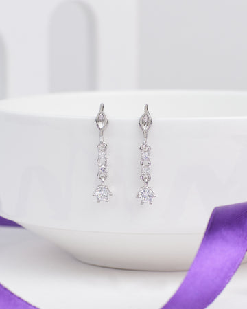 The Love Lite CZ Earrings by Chandrani Pearls are wearable silver drop earrings, each featuring three round clear CZ gemstones, shown hanging over a white bowl with a purple ribbon in the foreground.