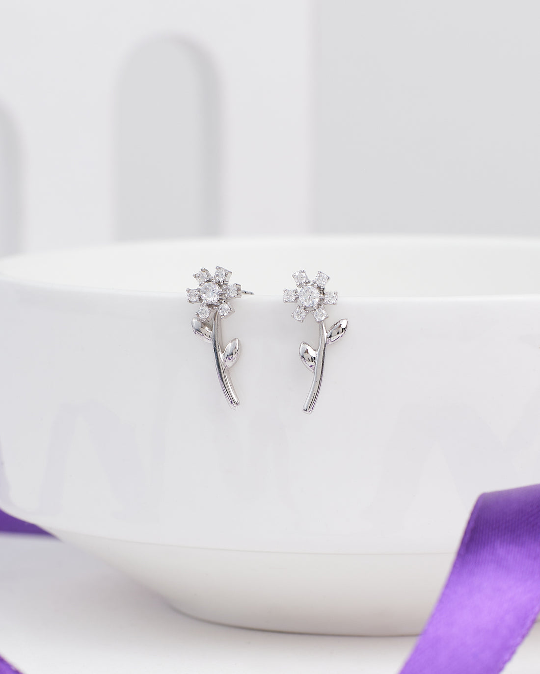 Glimmer Days Silver Studs by Chandrani Pearls—flower-shaped silver earrings with stems and leaves, ideal for daily wear—are shown resting on a white bowl, accented by a nearby purple ribbon.